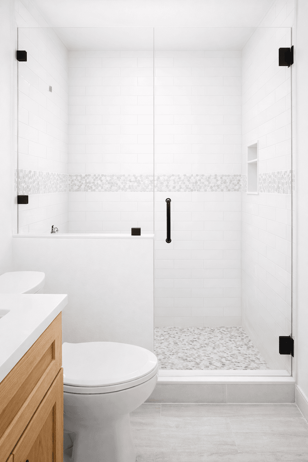 Frameless glass shower door with matte black hinges installed on newly tiled white subway tile with pebble mosaic accents